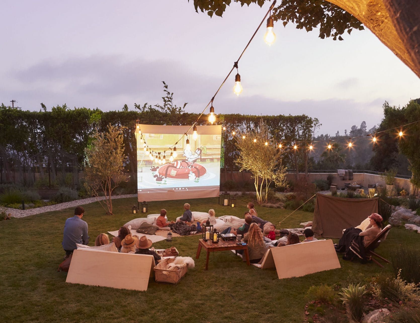 A group of people sitting on blankets in a garden with cartoons being played on an projector.