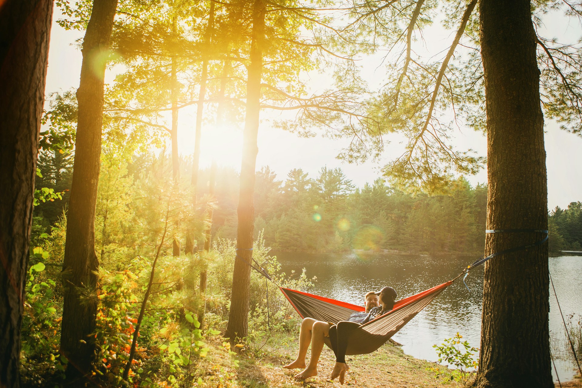 A couple sits in hammock in the woods by a lake. 