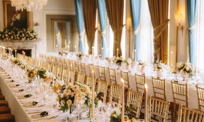 2 Intricately decorated long tables. Silver cutlery, tall white candles and white and green flower bouquets.