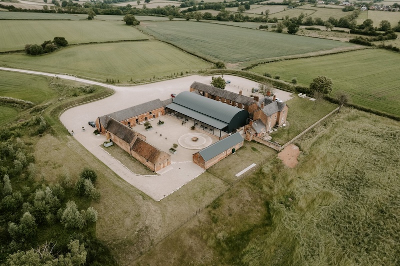 Aerial view of Grangefields Farm. Red brick barn conversion with central courtyard. Property surrounded by green grass fields. 