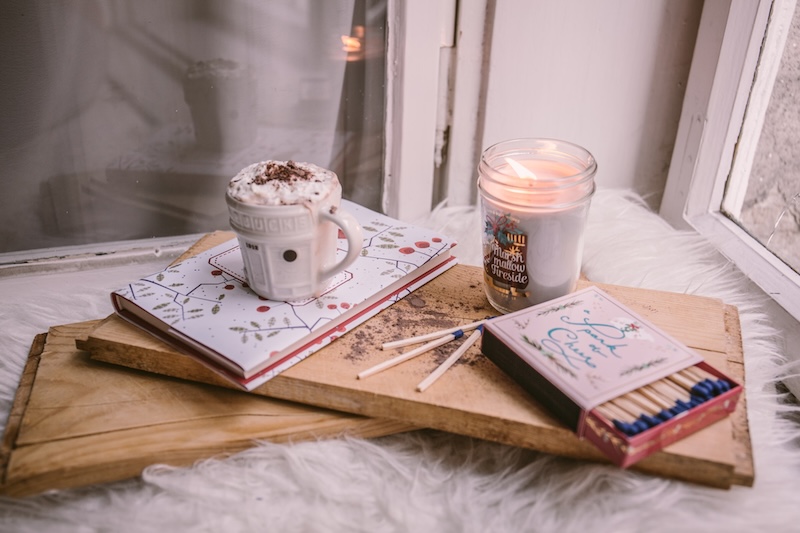 A book, hot chocolate, candle and matches a top a wooden board.