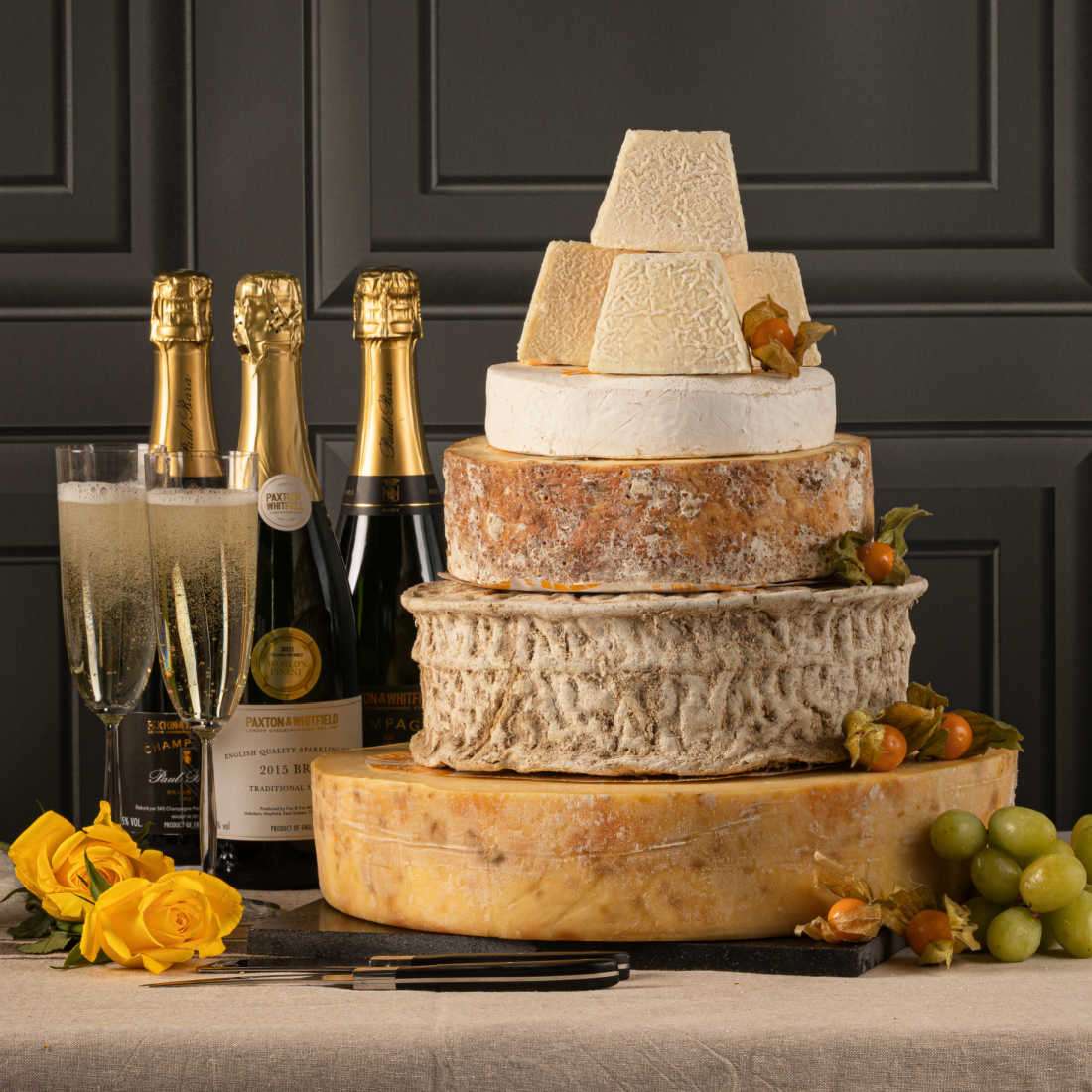 5 tiers of different cheeses emulating a wedding cake on a table with 3 bottle of champagne.