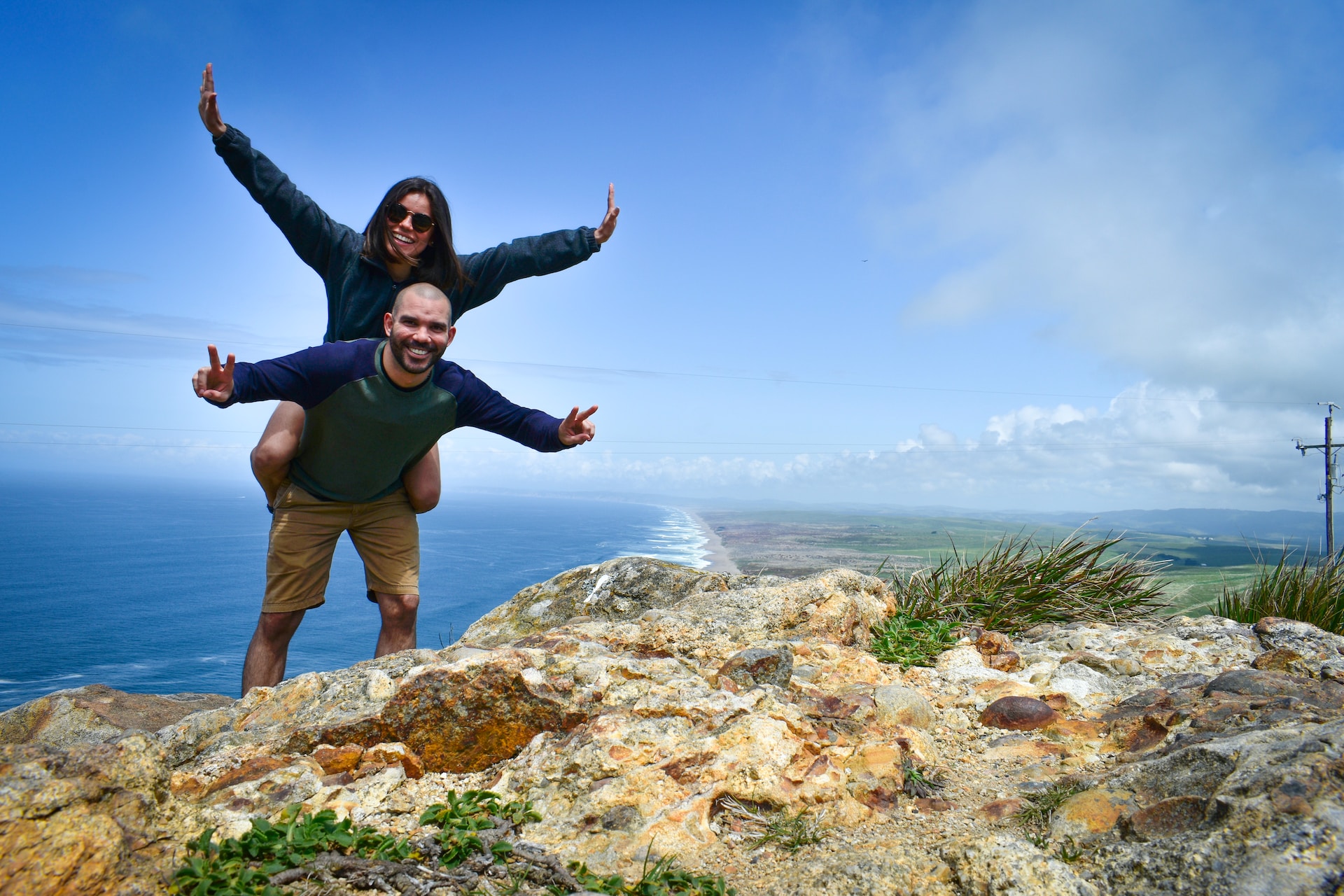 Wife poses on her husband's shoulders on top of cliff over looking the sea. 