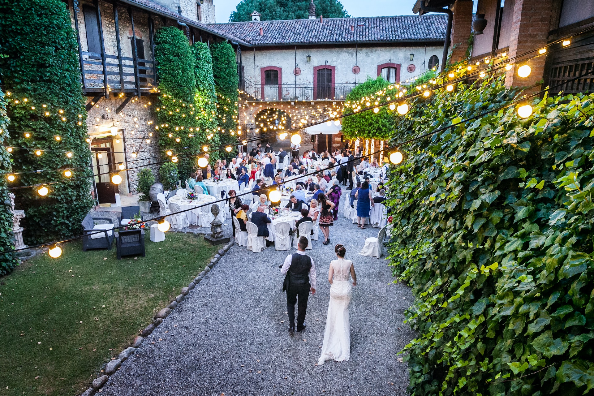 A bride and groom enter outdoor wedding reception under fairy lights. 
