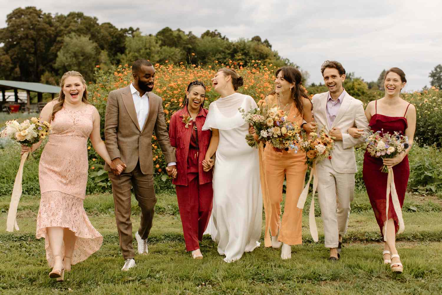 Wedding party of men and women walking through grass, laughing and smiling, holding bouquets of flowers. 