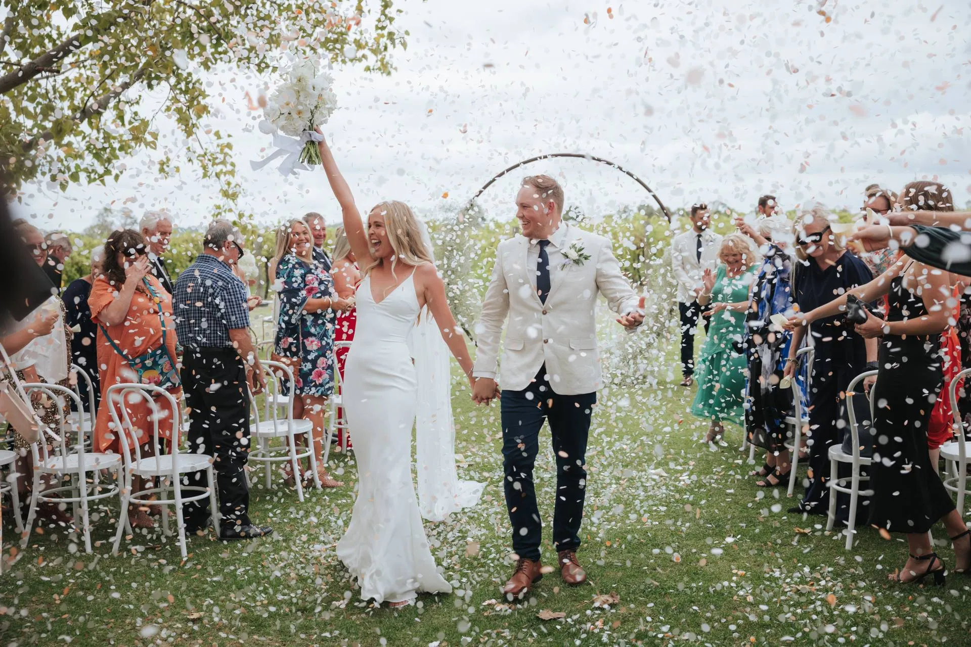Newly wed couple celebrate walking down outdoor aisle, with confetti being thrown over them. 